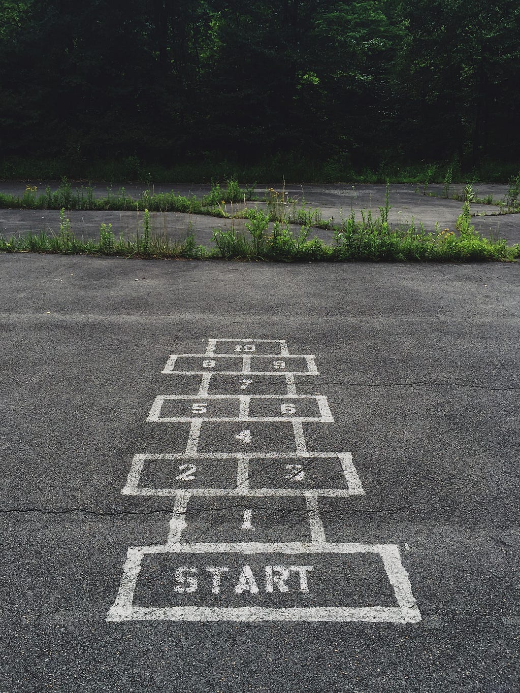 Photo of a mid grey tarmac space outdoors with a white outlines o a game of hopscotch, with a ‘start’ square in the foreground.