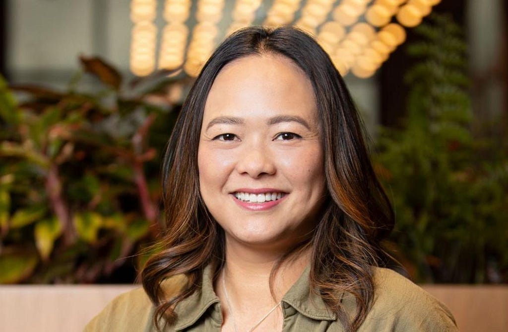 A headshot of Angela, looking at the camera and smiling with the background out of focus. She has shoulder-length dark brown hair and dark brown eyes. She is wearing a green button up shirt. She is seated indoors, in front of a row of plants.