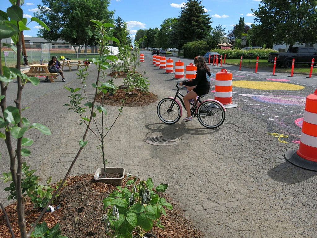 A girl rides her bike past a mural depicting stars and planets, painted on the asphalt, and between temporary planters with young trees.