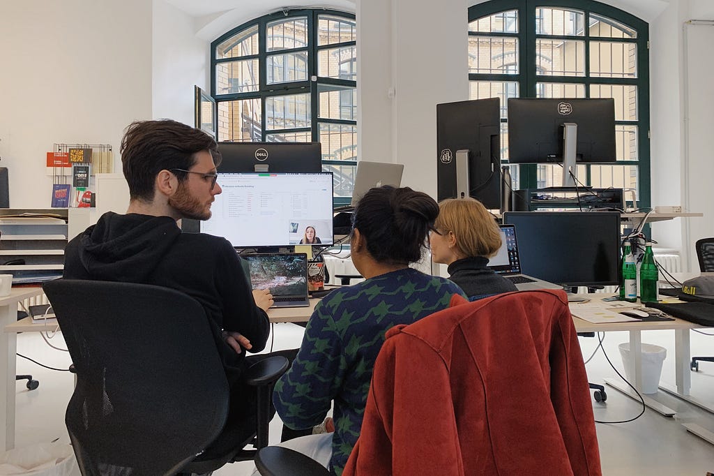 Three people sitting in front of a screen, discussing. On the screen is a fourth person connect via video call.