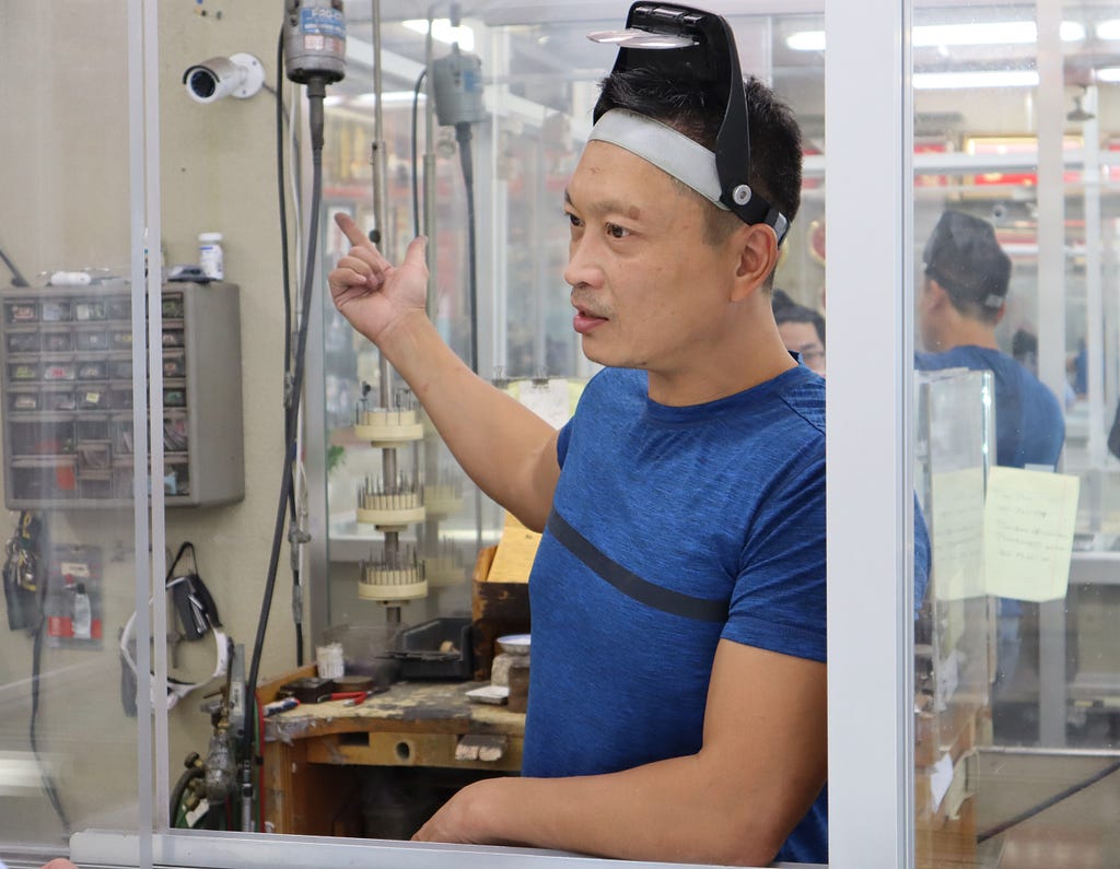 Phillip looks away from the camera, leaning on a glass case not visible to the viewer, mid-sentence, gesturing with his right hand behind him. He stands in front of his wooden work bench holding all the tools he uses to make jewelry. A mirror reflects his back in the background.