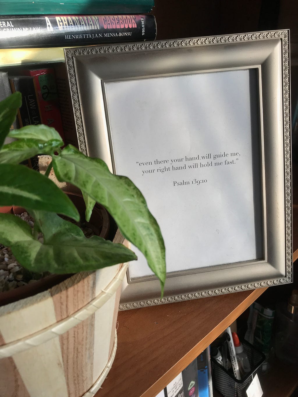 A gold-framed photo on a bookshelf. The inscription, “Psalm 139:10 — Even there, your right hand will guide me, your right hand will hold me fast,” is written in black text on white paper behind the glass. Adjacent to the frame is a criminal law textbook by Henrietta Mensah-Bonsu. To the right of the frame sits a potted arrowhead plant in a brown plastic pot, nestled within a wooden outer pot.