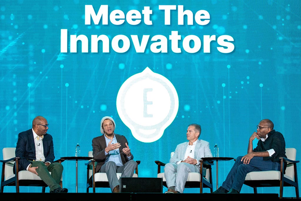 Photo of 4 panelists in business attire sitting in chairs on a stage. Behind them is a screen that says “Meet the innovators.”