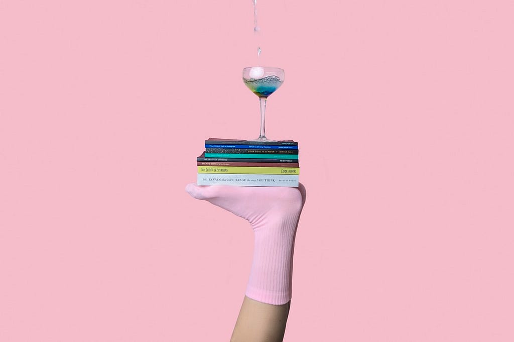 Women’s feet wearing candy-pink socks support a pile of books, on top of which a martini glass rests precariously