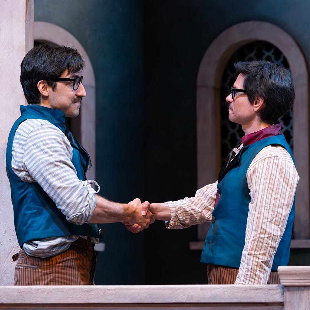 Two men dressed identically in blue vests, striped shirts, and black glasses shake hands and smile.