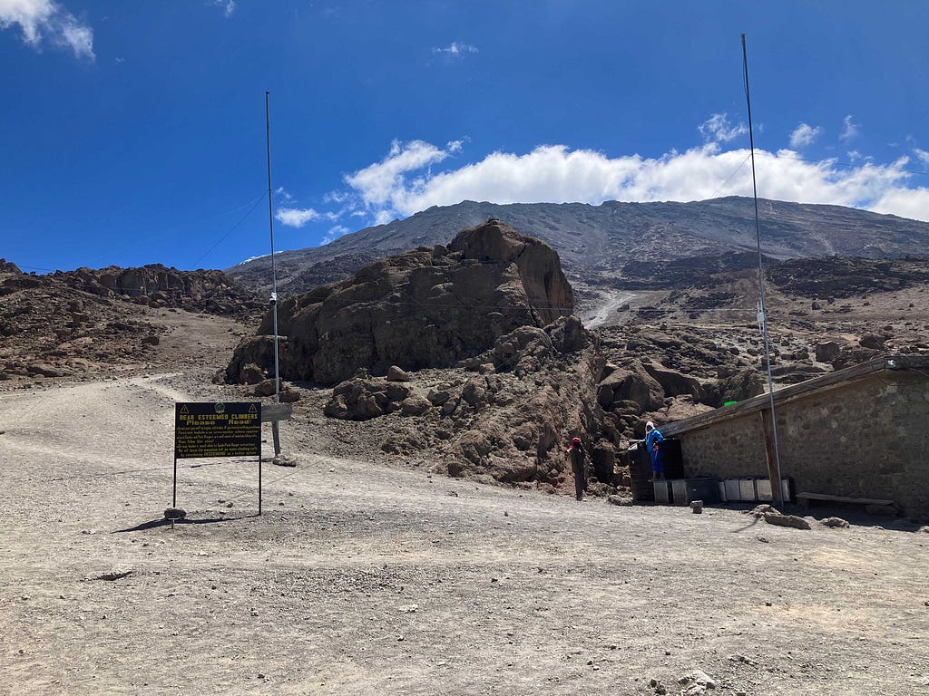 View from Kibo Huts looking toward the upper slopes of Mount Kilimanjaro, under blue skies with large boulders, a hut, and a sign in the foreground.