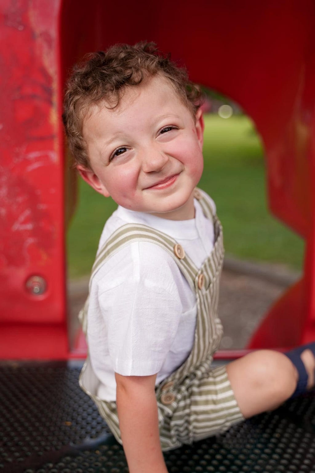 A young boy sitting on a bright red slide, smiling as he prepares to slide down.