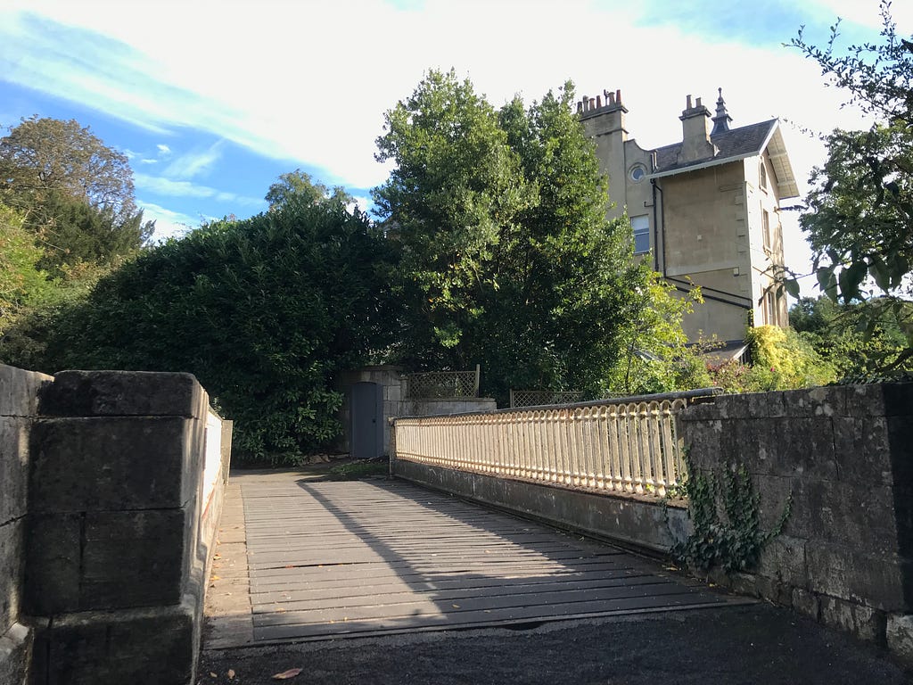 The bridge over the railway has low metal pillars on one side and they are cream. The bridge bottom is very flat and grey. You can see the trees the other side of the bridge — shrubberies and a house in the distance.