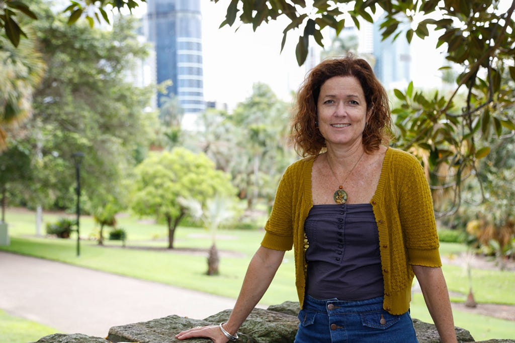 Photo of female researcher smiling for a photo, standing in front of a view of the Brisbane Botanical Gardens.