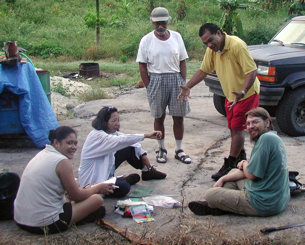 Ruth Utzurrum (second from left) doing field work in American Samoa. Ruth is sitting on the ground with two other people. There are two others standing.