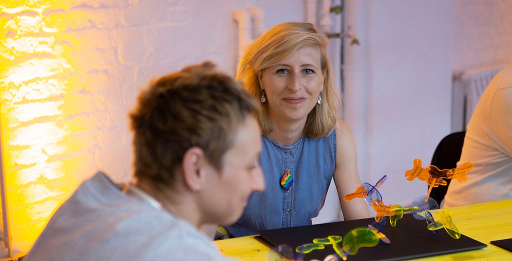 A photograph of a woman with blonde hair looking directly at the camera, smiling. In the foreground a table with colorful plastic pieces stacked together and a woman with short hair, not in focus.