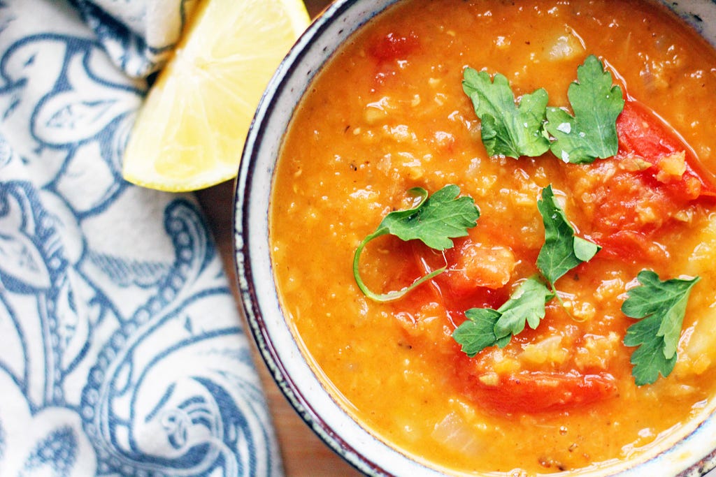 An overhead image of lentil and tomato soup garnished with cilanto next to a blue dishcloth and lemon wedge.