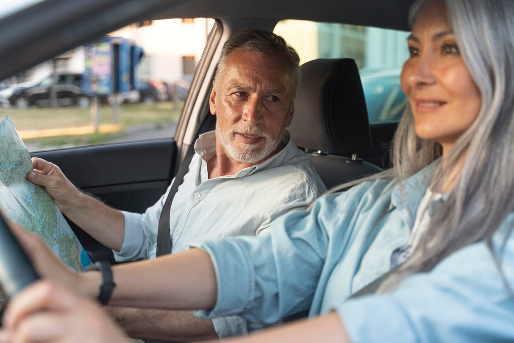 Two people are in a car, an older man and a younger woman. The man is holding a map and explaining something to the woman, who’s driving with a frozen sort of smile on her face.