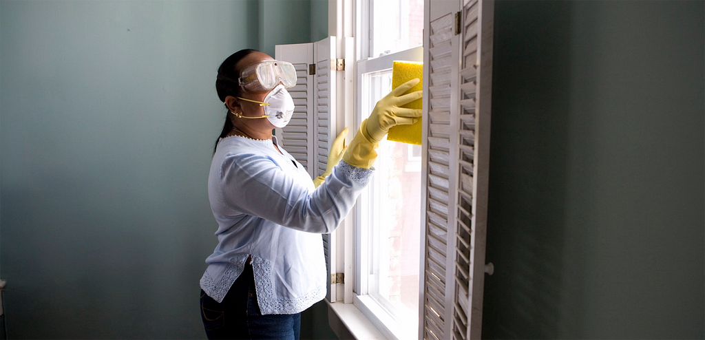 a woman with goggles, mask, and yellow gloves cleans a window with a sponge