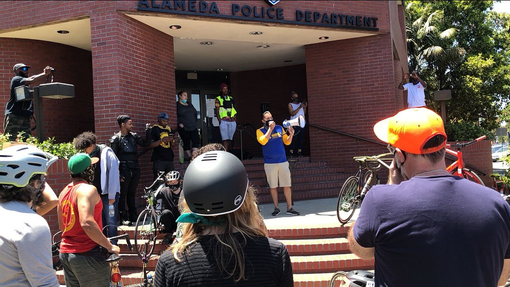 A group stands outside the Alameda Police Department building, one person in the center holds a megaphone, talking to the group