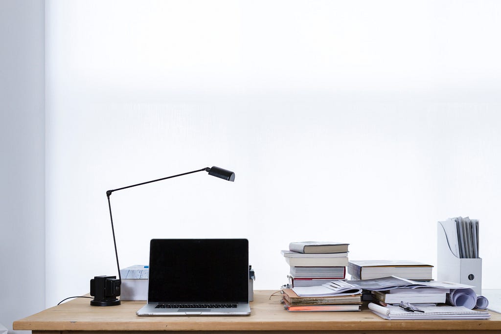 Minimalist white-background photo of a natural wood remote work desk with black pendant lamp, laptop, piles of books, and various papers mixed with writing implements and a file case