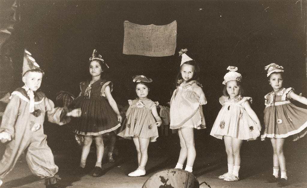 A group of six children who are about four to six years old on a stage. Five girls wearing hats hold their dresses out to their sides as if curtsying. A boy in a clown costume at far left is about to walk in front of the girls.