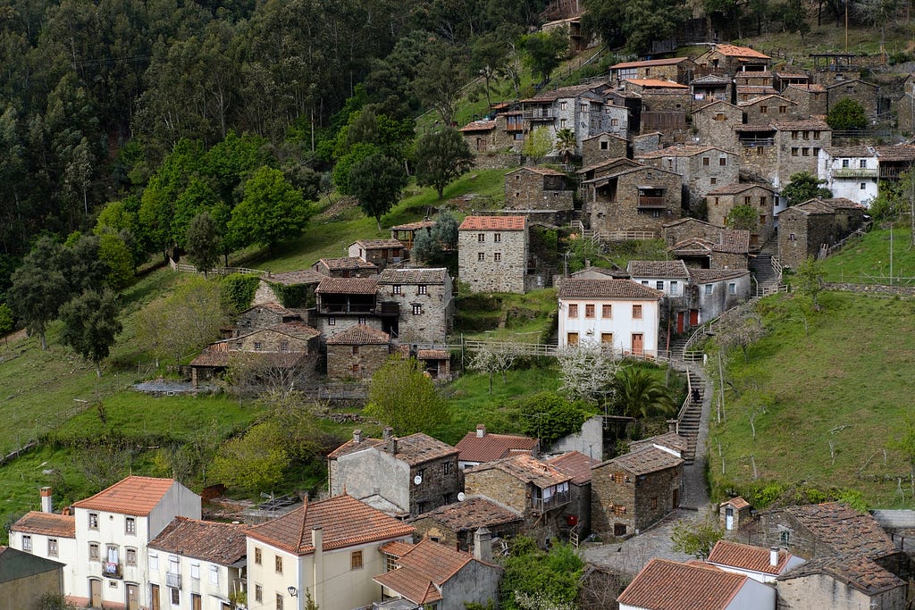 An aerial shot of a village — Candal, Lousã, in Portugal. Among the very green grass and trees, the many houses are built along the natural slope of the mountain, some of newer materials, and some of far more antiquated origins.