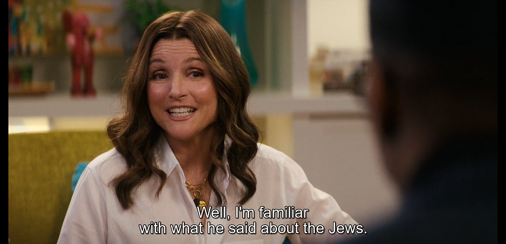 A woman with a fake smile is speaking. Subtitles read “Well, I’m familiar with what he said about the Jews.”