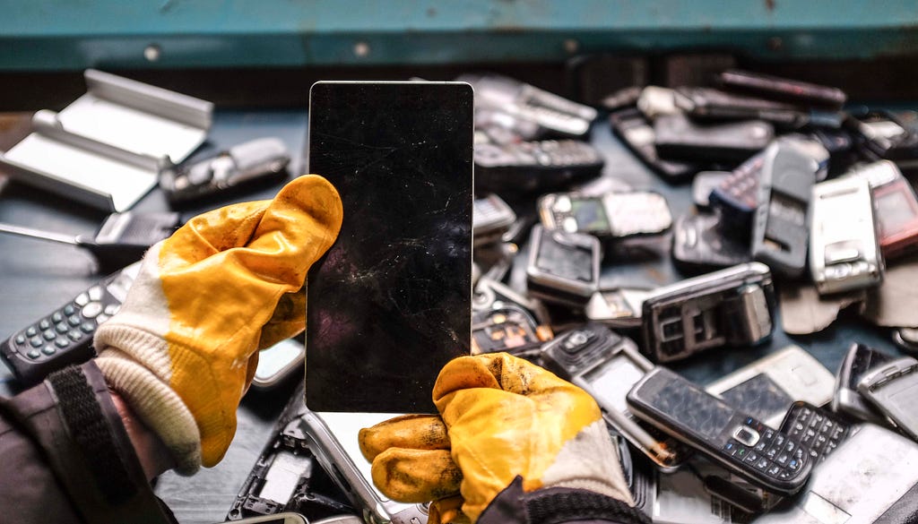 Someone wearing gloves holding a smart phone amid lots of old and broken mobile phones.