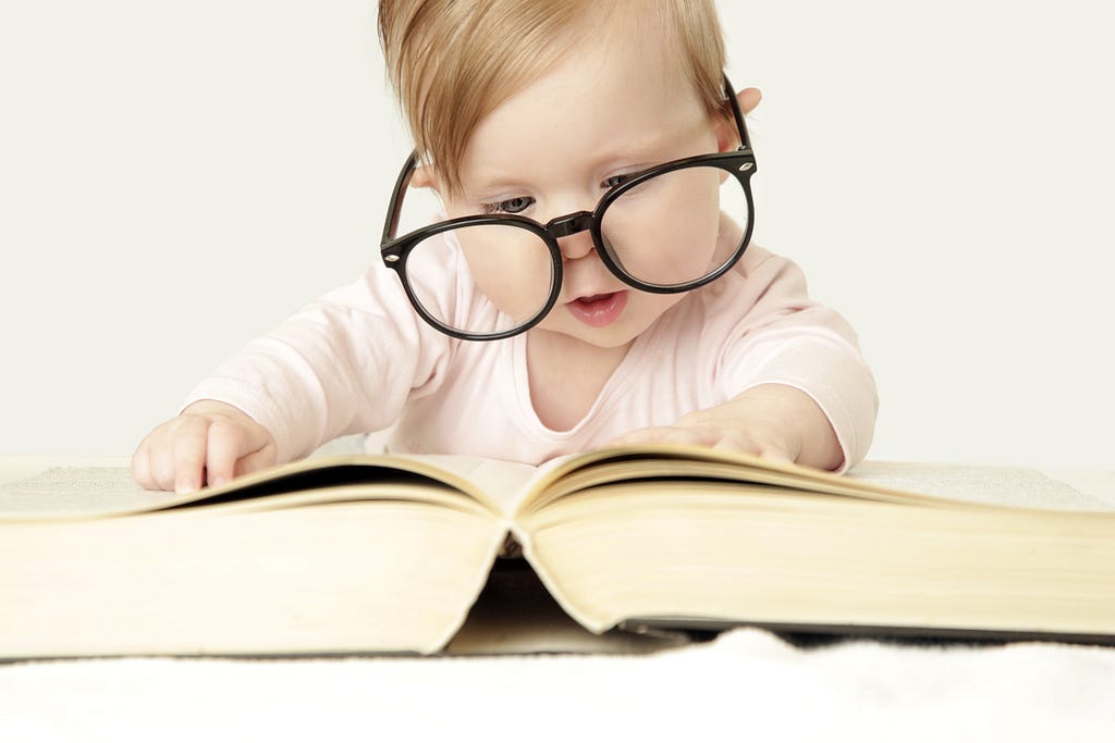 Copil de un an cu ochelari de adult în fața unei cărți groase