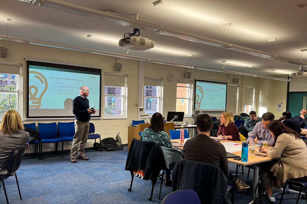 An image showing Professor Stuart Taberner addressing the first Horizons Institute collaborative lab. There are a number of people sat listening around groups of tables, and there is a powerpoint screen in the background.