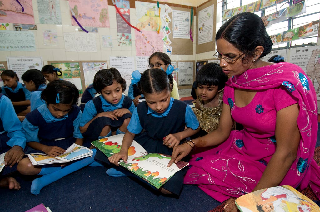 A female teacher in a pink Indian dress teaching Indian girls how to read a picture book