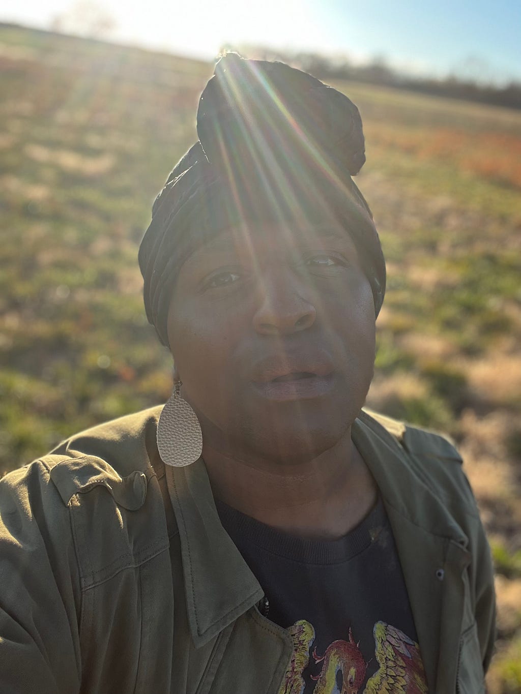 Black trans human standing in a field with the sun on their back. the human is breathless and knowing.