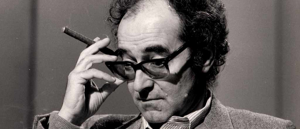 Black and white photo of what looks like Peter Sellers but is in fact Jean Luc Godard peering over his glasses while holding a cigar.
