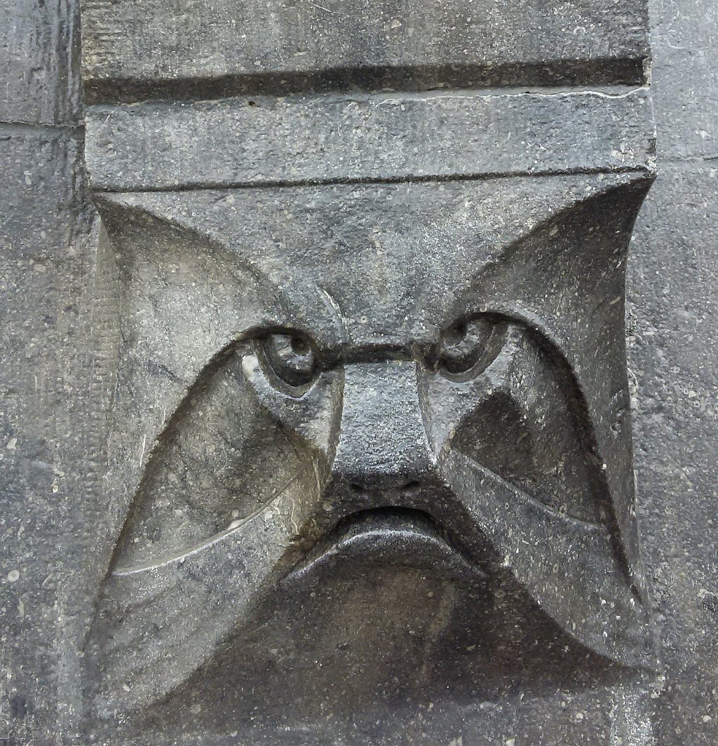 A frightening stern face, a mascaron, carved in gun-metal gray stone peers out from a doorway in Amsterdam. Photo by author.