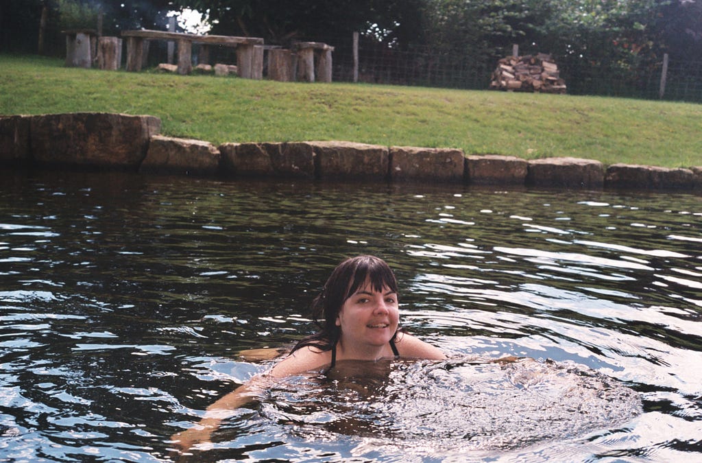 Anna swims in lake, smiling, in grassy background with benches round a fire pit in Sussex, UK