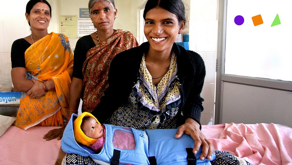 A foto apresenta três mulheres e uma delas carregando um bebê que está dentro de uma manta azul