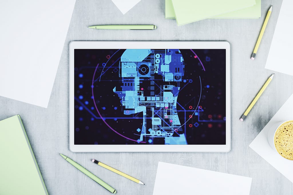 Top-down view of a tablet on a grey desk displaying a digital illustration of a human head profile composed of circuit board patterns and mechanical elements in blue tones against a dark background. Yellow and green pencils, sticky notes, papers, and a cup of coffee surround the tablet.