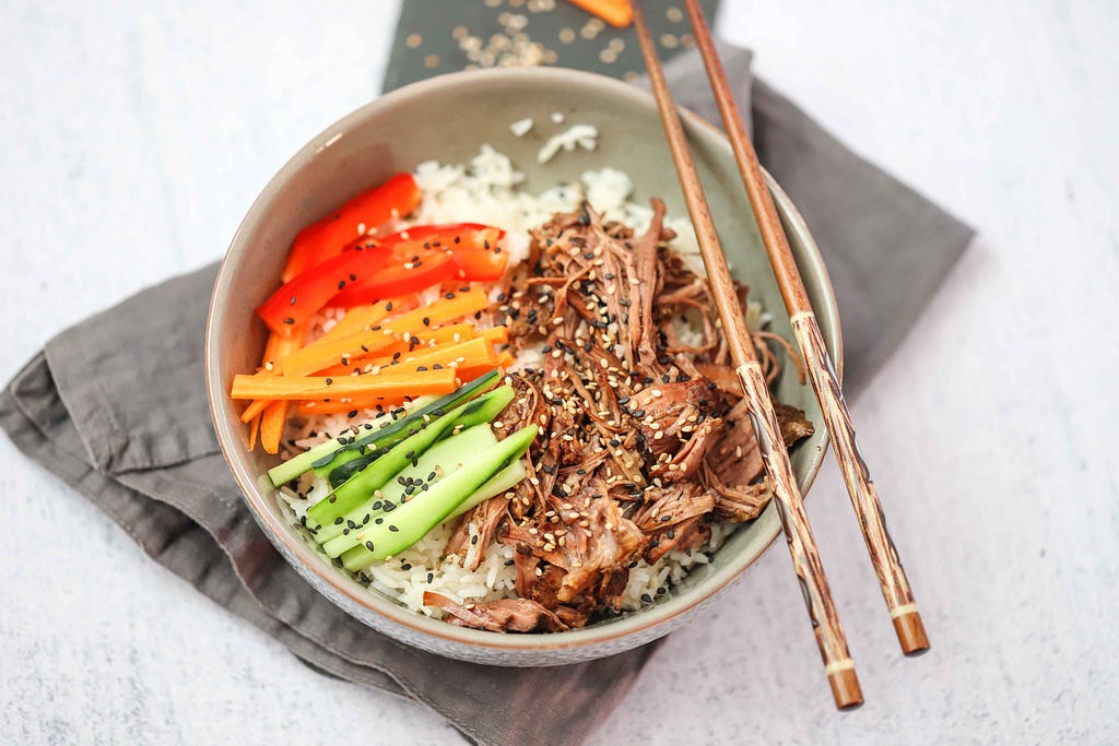 A bowl of slow cooked honey garlic beef alongside cucumbers, carrots, and red bell pepper.