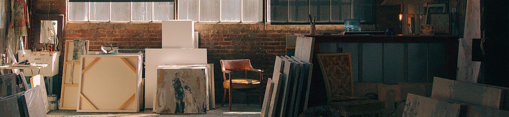 Painted canvases are leaned against a brick wall beneath wide multi-paned windows.