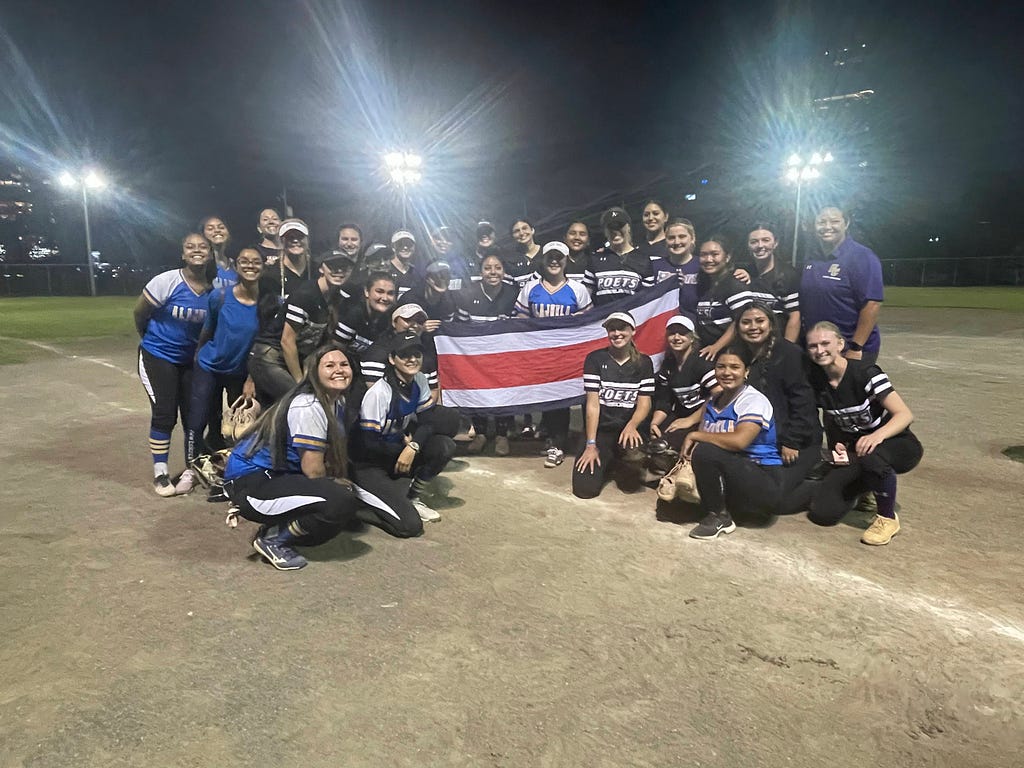 Photo of the Whittier Softball team alongside the team in Costa Rica, holding the Costa Rican flag on the softball diamond.