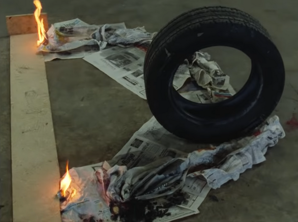 A tire sits on the tip of a v-shaped pile of newspaper. 2 flames on either side are just beginning to ignite the newspaper.