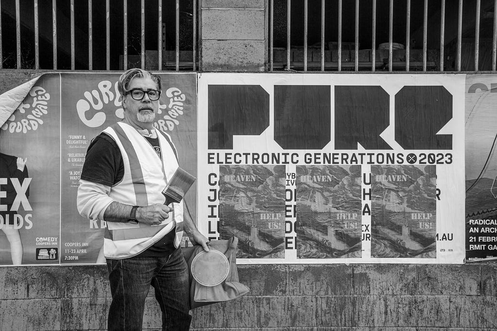 Richard Harding stand outside in a hi-vis vest, holding a paintbrush. Behind him, his past-ups have an image of a submarine with the words ‘heaven help us’.