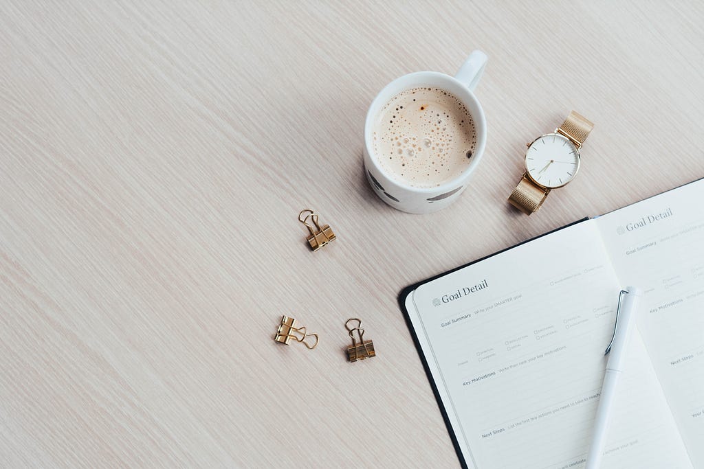 A cup of coffee, a watch, paper clips, and a notebook showing a list of goals with a pen placed on it — an illustration of habit planning at the beginning of the year.