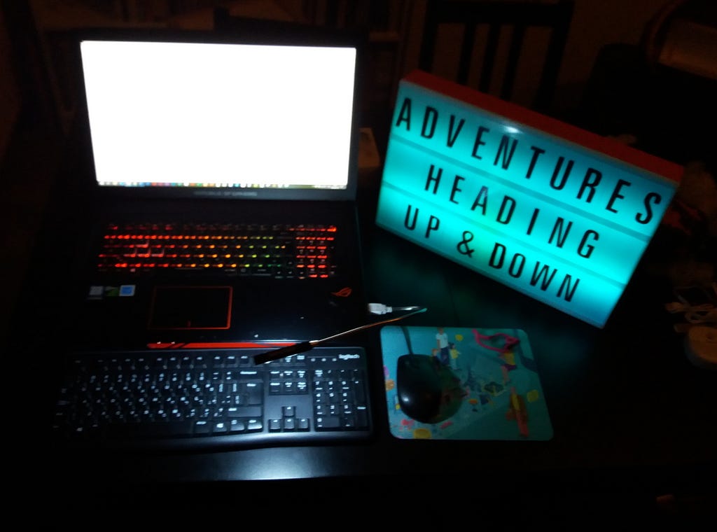 A blank, white laptop screen shining in the dark, and a lightbox with the words ‘adventures heading up and down’.