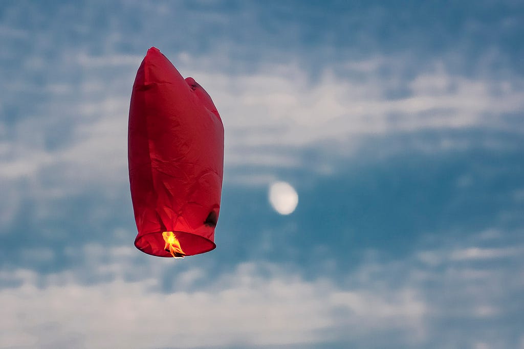 Photograph of a small hot-air propelled balloon-type thingie floating in the sky with an out-of-focus gibbous moon in the background.