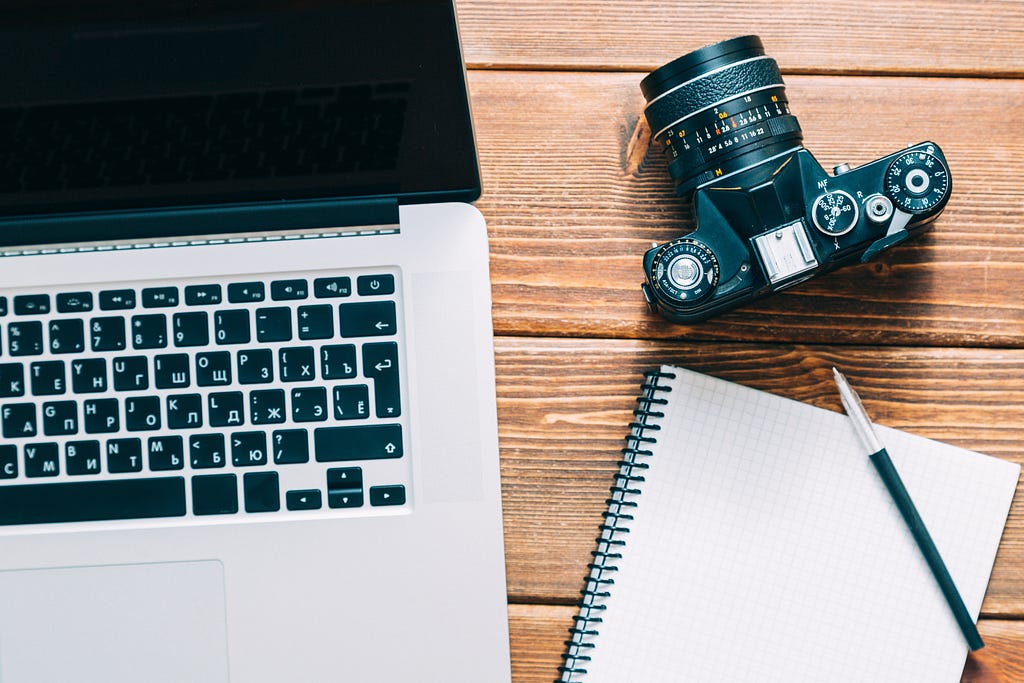 Flatlay image of a desk with a laptop, notebook, and camera arranged neatly on it.