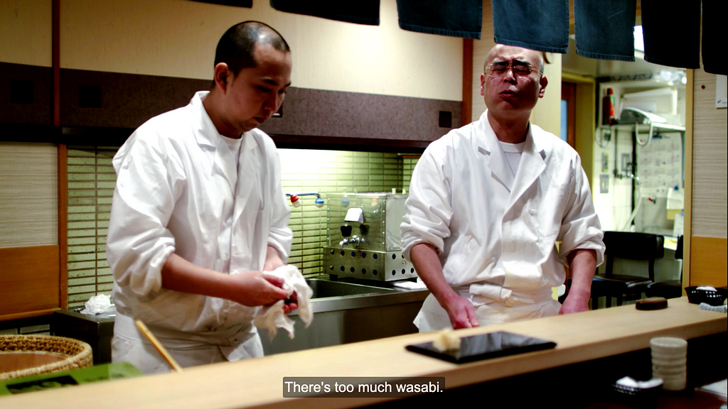 Two chefs standing up in a kitchen. The older chef pulls a face as he tries the younger chef’s sushi