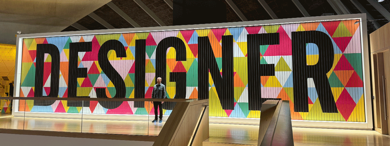 Animated gif of Lewis standing infront of a large multicoloured sign which rotates through the words ‘Designer’, ‘Maker’, and ‘User’.