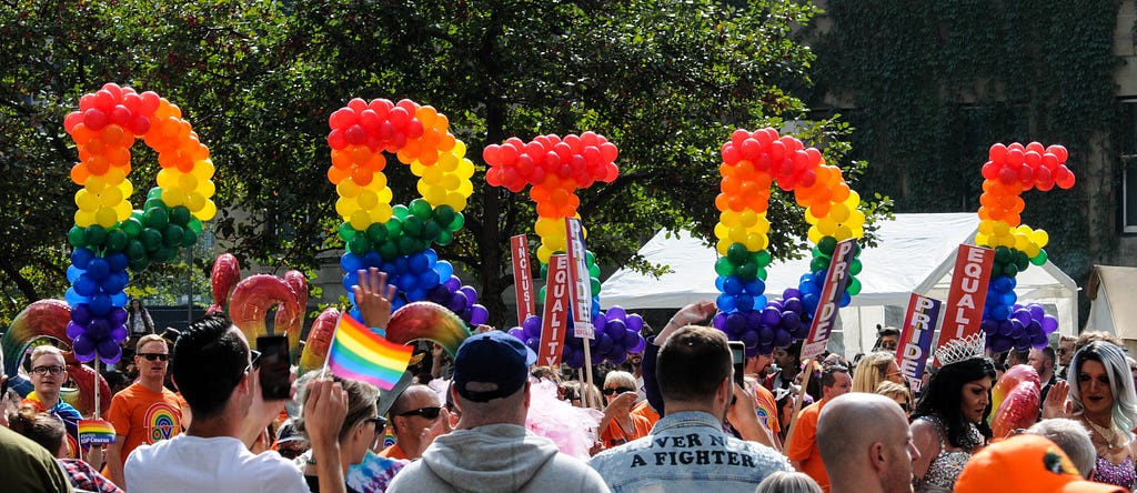 A photo of a crowd of people with pride flags and a sign spelling PRIDE in front of them created out of balloons in rainbow order