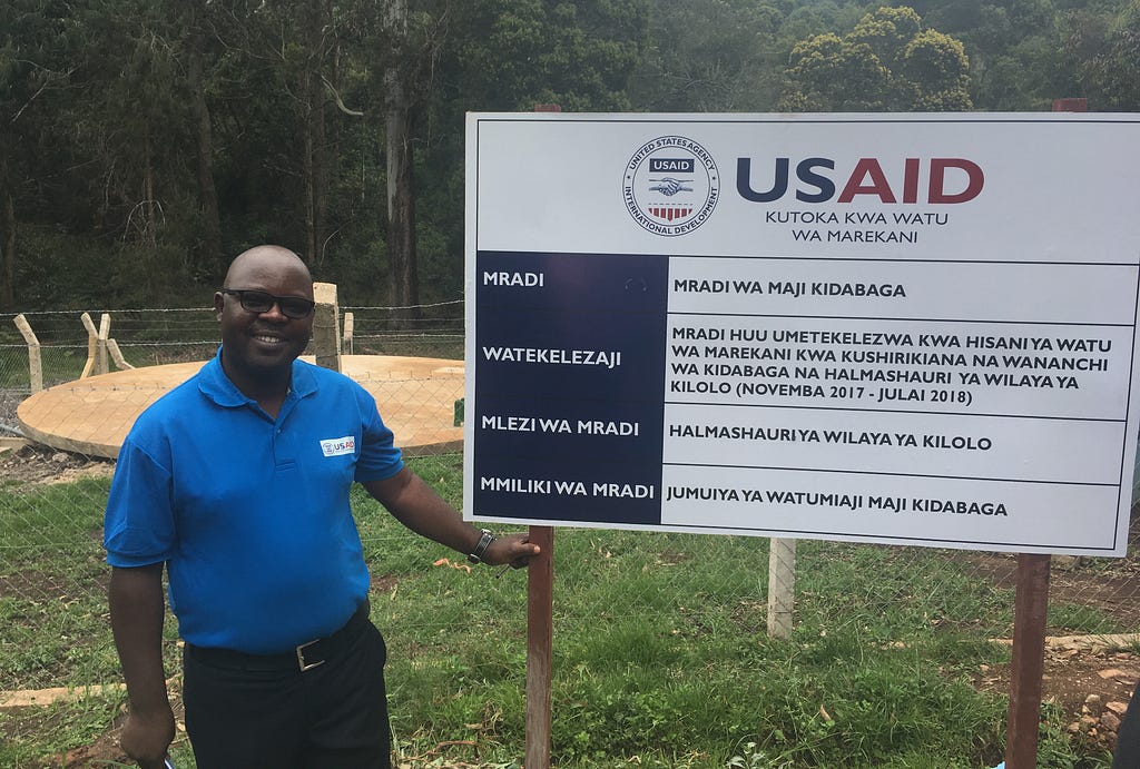 A man stands outside next to a small billboard describing USAID’s Maji Na Usafi Wa Mazingira activity in Tanzania.