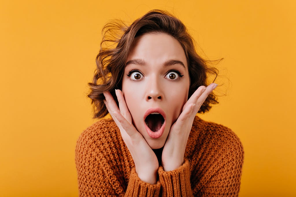 A girl in an orange sweater pressing her hands to her face and looking shocked and amazed.