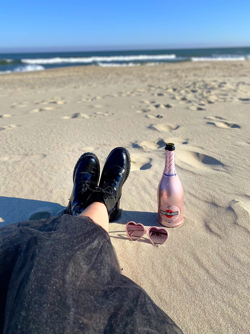 A pink bottle of champagne, heart-shaped sunglasses and a girl’s feet in combat boots on the beach.