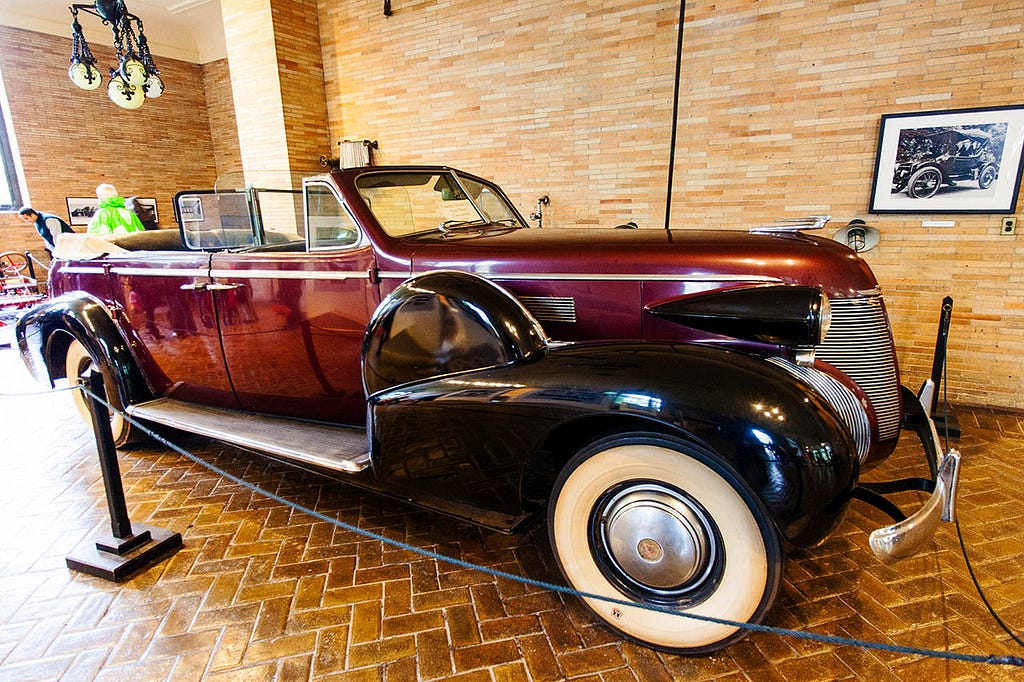 A classic car, possibly a Cadillac Series 70 from the 1930s, on display in a museum with a brick wall background. The car is red with black fenders and has a chrome grille.