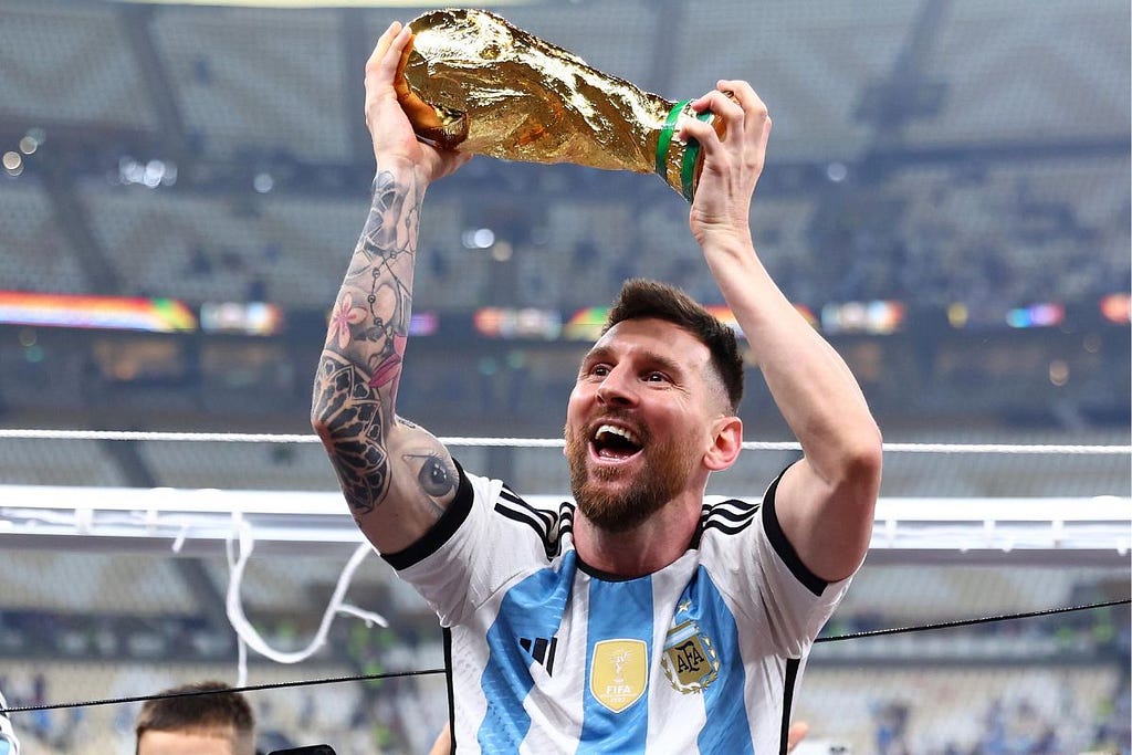 Messi sorrindo de boca aberta erguendo a taça da Copa do Mundo com as duas mãos com o estádio ao fundo